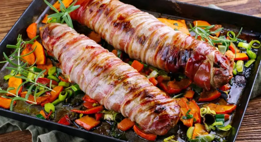 Dwie polędwiczki wieprzowe zawinięte w boczek, pieczone z warzywami. Idealny sposób, jak zrobić polędwiczki wieprzowe, żeby były miękkie i soczyste.