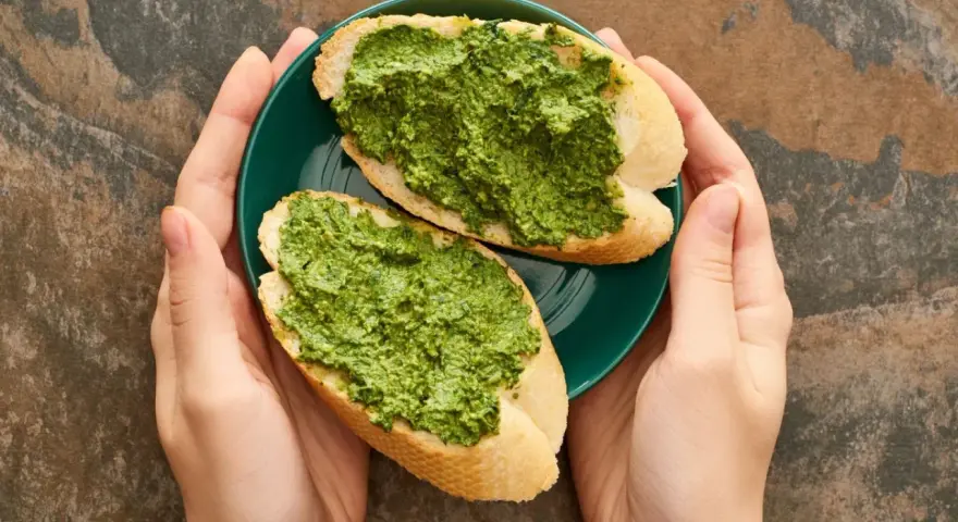 Dwie kromki bagietki z aromatycznym pesto z pietruszki na zielonym talerzyku, trzymanym w dłoniach.